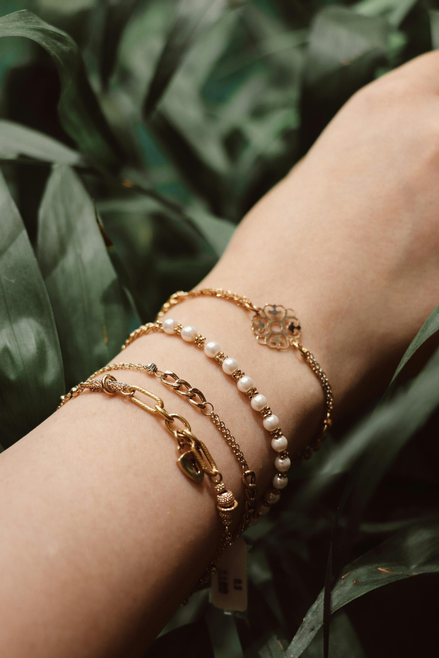 Close-up of gold and pearl bracelets on a wrist amidst green leaves for a natural, elegant look.